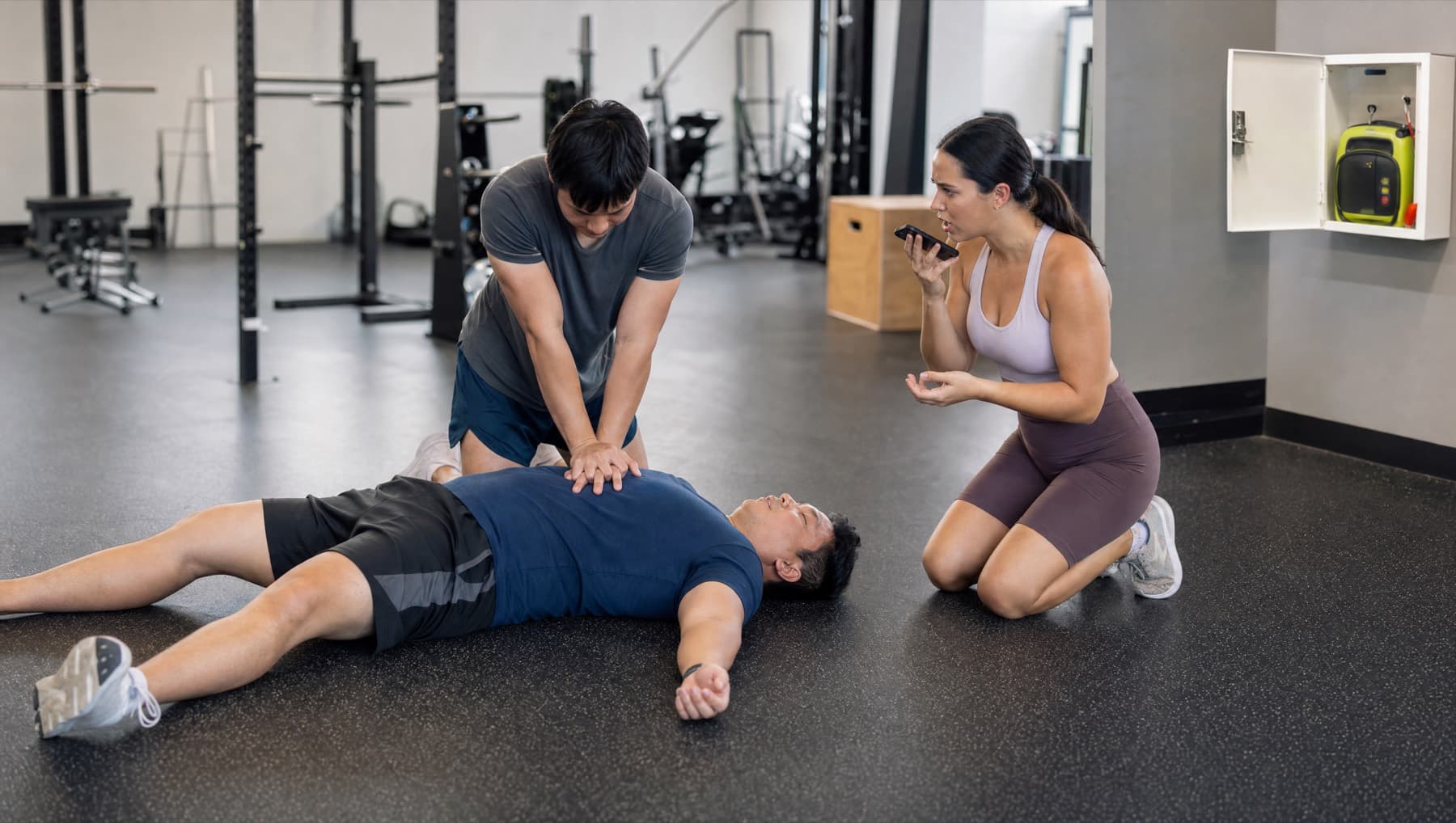 A gym member gives chest compressions to a collapsed man while another calls for help, with a wall defibrillator cabinet open
